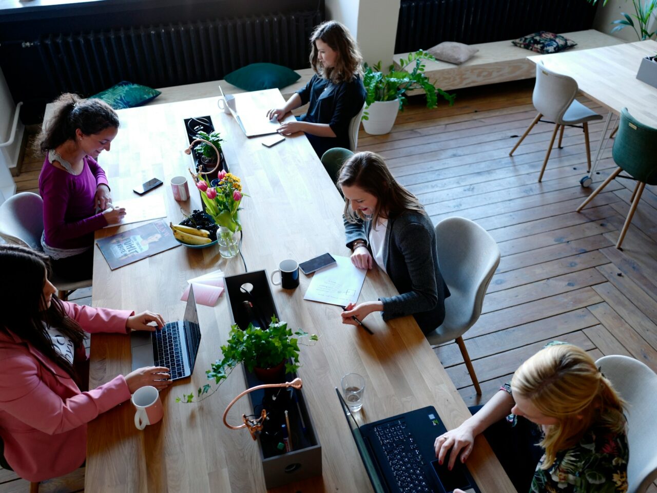 Five women working at a large wooden table in a bright office space. Each is using a laptop, with notebooks and plants on the table. There are casual seating areas in the background.