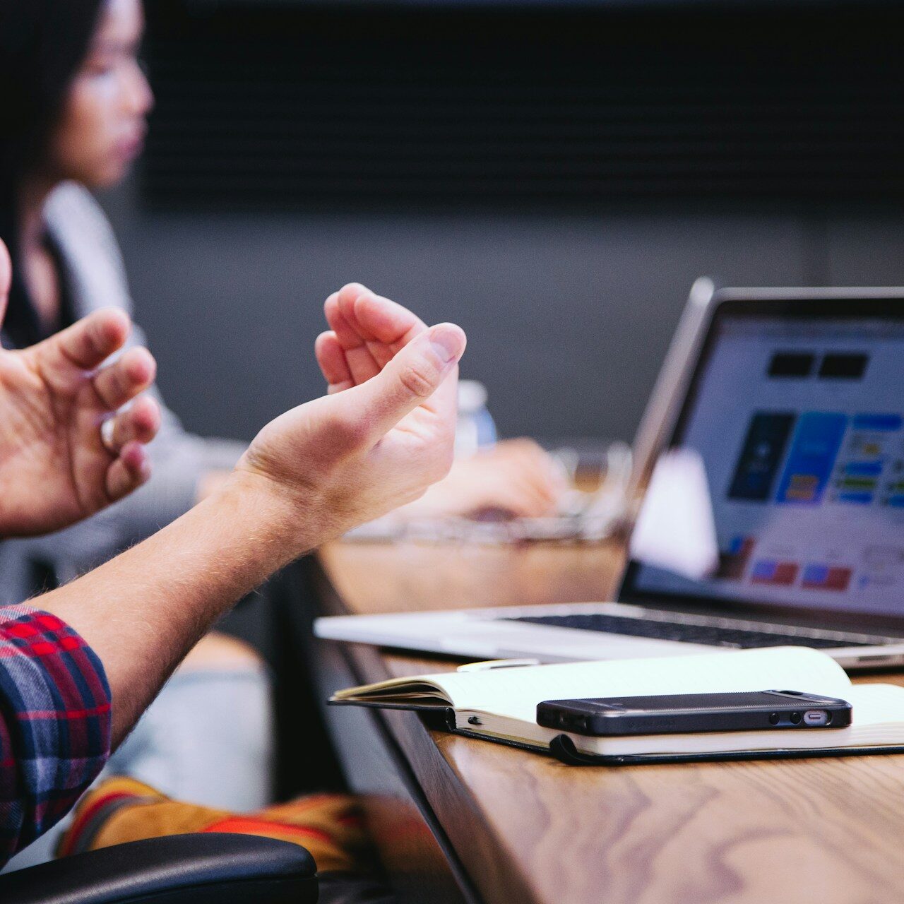 headway-5QgIuuBxKwM-unsplash A close-up shot of a person's hands gesturing during a discussion in a meeting, with a laptop displaying design options on a table. A woman is seated in the background, partially visible.