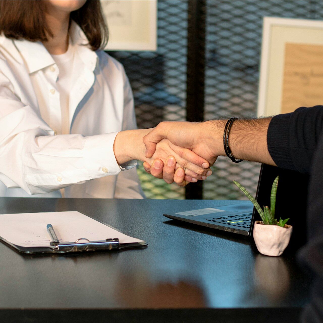 A close-up of two people shaking hands across a black desk, one wearing a white shirt and the other in a dark sweater. On the desk, there is a clipboard with a pen and a small potted plant beside a laptop.
