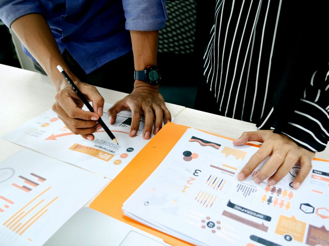 Two individuals working collaboratively at a desk, reviewing printed documents with graphs and charts. One person is writing notes while the other is examining the materials. A laptop is visible in the background.