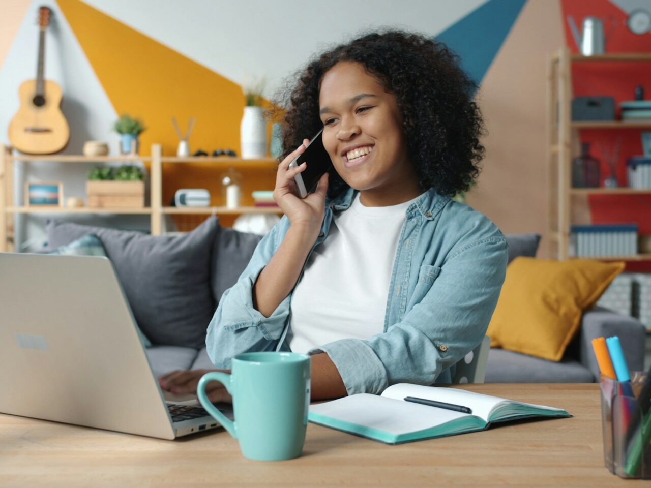 A woman with curly hair is sitting at a desk talking on a phone while working on a laptop. A teal mug and a notebook are on the desk, with various colored pens beside them. The background features colorful geometric wall designs and shelves with items.